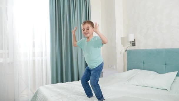Slow motion van vrolijke lachende jongen springen en dansen op groot bed in slaapkamer met groot raam — Stockvideo