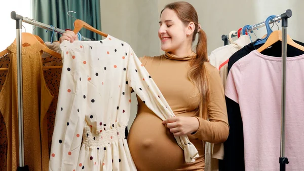 Smiling pregnant woman taking beautiful dress hanging on hanger from clothes rack at her home — Stock Photo, Image