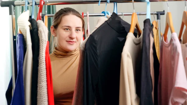 Beautiful smiling woman behind long rack of clothes on hangers choosing dress to wear — Stock Photo, Image