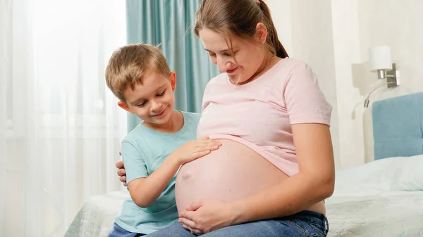 妊娠中の母親の大きな腹に触れるかわいい男の子がベッドルームに座っています。赤ちゃんを期待している家族の肯定的な感情 — ストック写真