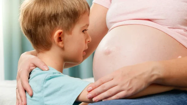 Portret van kleine zoon praten en luisteren naar grote zwangere moeder buik in de slaapkamer — Stockfoto