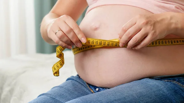 Крупный план беременной женщины, сидящей на кровати и измеряющей свой большой живот измерительной лентой — стоковое фото