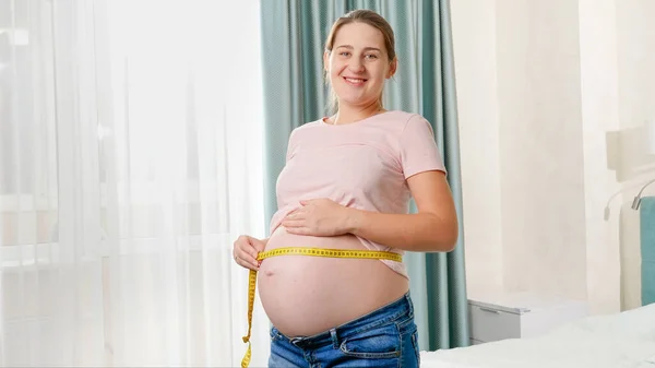 Feliz mujer embarazada sonriente de pie en la ventana del dormitorio y midiendo el vientre grande con cinta métrica — Foto de Stock