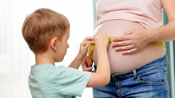 Küçük çocuk hamile annesine yardım ediyor. Büyük göbeğini bant ile ölçüyor. Hamilelik süresince boyut ve vücut parametreleri kontrol ediliyor — Stok fotoğraf