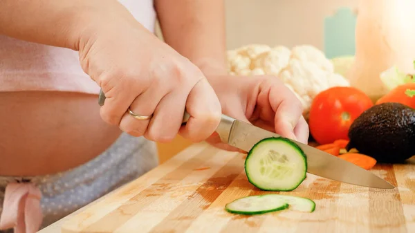 Close-up van zwangere vrouw koken op keuken en het snijden van komkommers met scherp mes op houten snijplank — Stockfoto