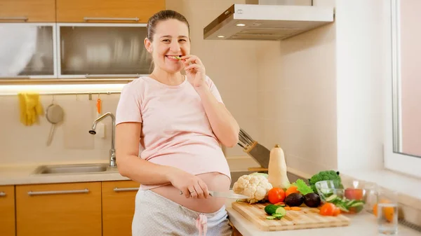 Porträt einer glücklich lächelnden Schwangeren, die in der Küche kocht und frisches, schmackhaftes Gemüse isst. Konzept einer gesunden Lebensweise und Ernährung während der Schwangerschaft — Stockfoto