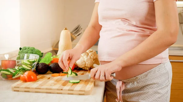 Улыбаясь беременной вумен делает салат и ест свежий огурец на кухне в доме. Концепция здорового образа жизни и питания во время беременности — стоковое фото