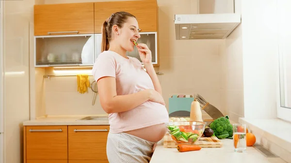Mujer embarazada sonriente esperando al bebé comiendo verduras frescas y mirando por la ventana de la cocina. Concepto de estilo de vida saludable y nutrición durante el embarazo — Foto de Stock