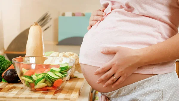 Молода вагітна жінка готує і їсть овочевий салат, який тримає великий живіт і торкається його руками — стокове фото