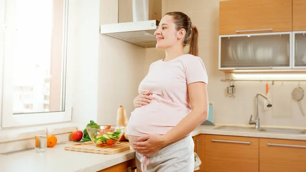 Sourire jeune femme enceinte toucher et tenir gros ventre regardant par la fenêtre sur la grande cuisine.Concept de mode de vie sain et la nutrition pendant la grossesse — Photo