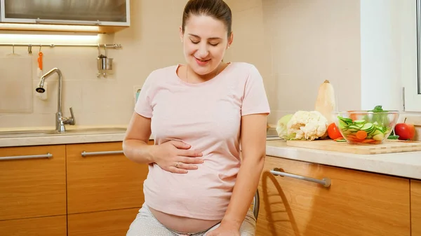 Retrato de la mujer embarazada sonriente sentada en la silla en la cocina después de hacer las tareas domésticas y cocinar —  Fotos de Stock