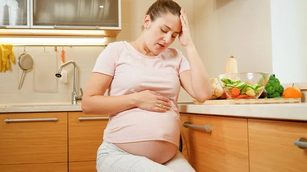 Mujer embarazada joven con dolor en el vientre que descansa en la silla después de cocinar y trabajar en la cocina —  Fotos de Stock