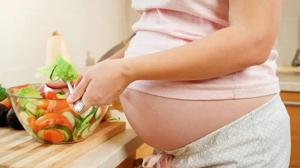 Close-up van zwangere jonge vrouw die groenten mengt in verse smakelijke salade op de keuken. Concept van gezonde levensstijl en voeding tijdens de zwangerschap. — Stockfoto