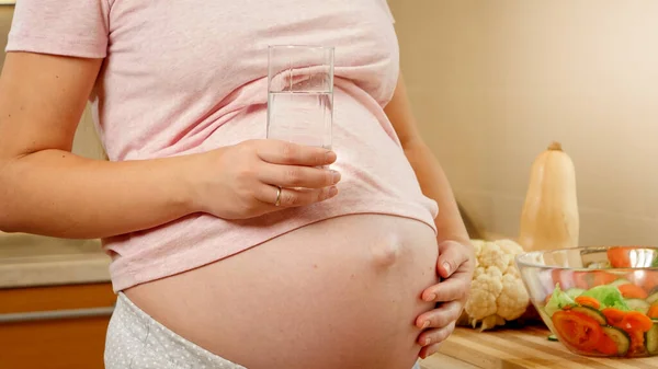 Karnında bir bardak suyla mutfakta duran hamile kadının yakın çekimi. Hamilelik süresince sağlıklı yaşam tarzı ve beslenme kavramı — Stok fotoğraf