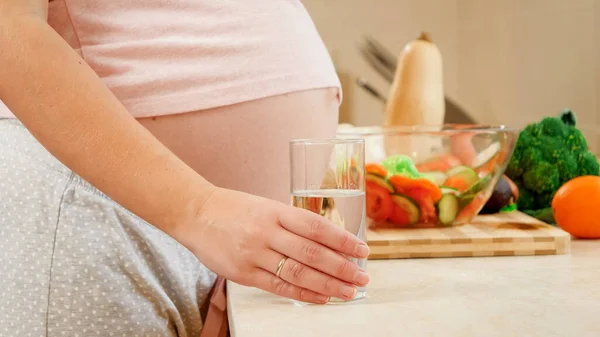 妊娠中の女性のクローズアップは、台所のテーブルの上から水のガラスを取って赤ちゃんを期待。妊娠中の健康的なライフスタイル、栄養と水分補給の概念 — ストック写真