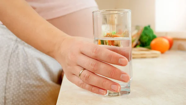 妊娠中の女性の閉鎖は、台所のテーブルの上から澄んだ水のガラスを取ります — ストック写真
