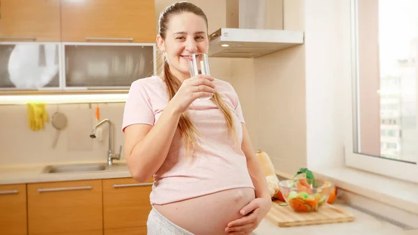 Glad gravid kvinna dricksvatten på köket på morgonen och ler mot kameran — Stockfoto