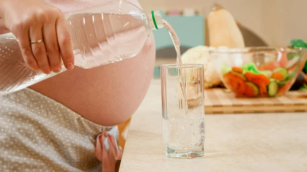 Closeup of pregnant woman pouring water in glass from plastic bottle on kitchen at morning. Concept of healthy lifestyle, nutrition and hydration during pregnancy — Stock Photo, Image