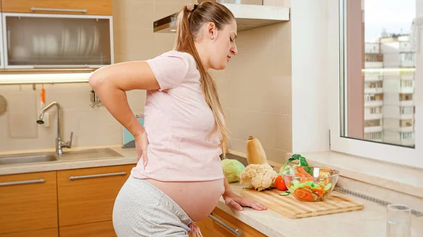 Mujer embarazada joven que sufre de dolor espinal cogida de la mano en la espalda y descansando en la cocina después de cocinar y hacer las tareas domésticas. —  Fotos de Stock