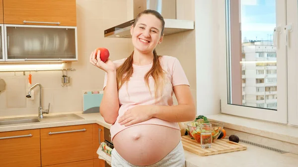 Glad leende gravid kvinna har kul med rött moget äpple på köket på morgonen. Begreppet hälsosam livsstil, kost och hydrering under graviditeten — Stockfoto