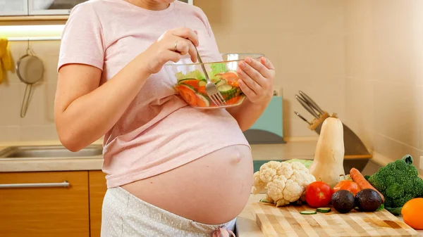 Primer plano de la mujer embarazada con el vientre grande sosteniendo tazón de vidrio con ensalada de verduras y comer en la cocina Imágenes de stock libres de derechos