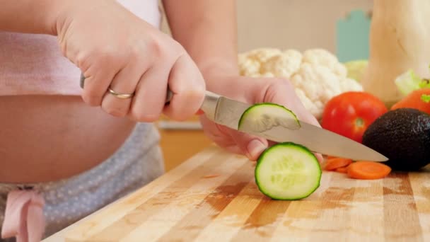 Крупный план приготовления пищи беременной женщиной на кухне и резки огурцов острым ножом на деревянной доске — стоковое видео