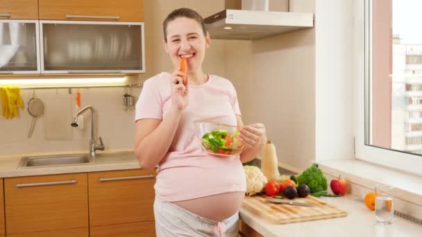 Felice donna incinta sorridente con grande pancia che tiene grande ciotola di insalata vegetale e posa in cucina con grande finestra — Video Stock