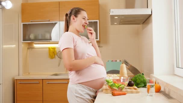 Sorridente donna incinta in attesa di bambino mangiare verdure fresche e guardando fuori dalla finestra sulla cucina. Concetto di stile di vita sano e nutrizione durante la gravidanza — Video Stock
