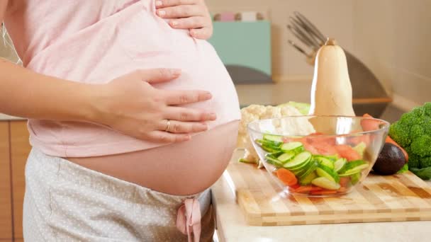 Крупный план беременной женщины с большим животом, стоящей на кухне и трогающей большой живот. Концепция здорового образа жизни и питания во время беременности — стоковое видео