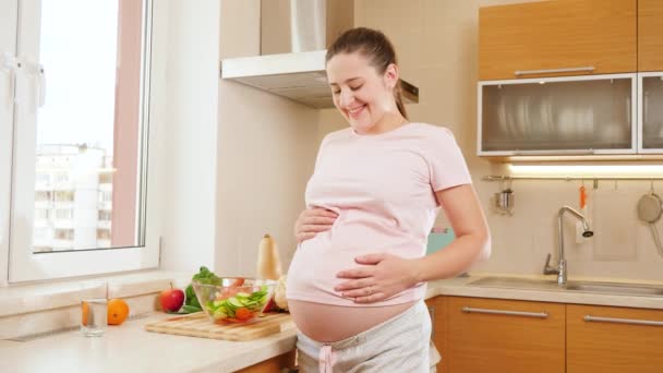 Sorridente giovane donna incinta toccando e tenendo grande addome guardando fuori dalla finestra sulla grande cucina.Concetto di stile di vita sano e nutrizione durante la gravidanza — Video Stock