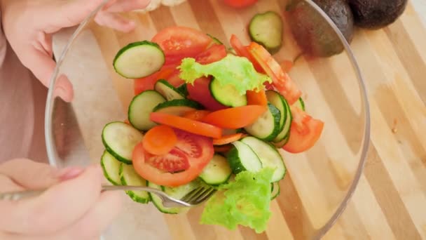 Вид зверху на жінку, змішування та перемішування нарізаних свіжих овочевих скибочок у смачному салаті у великій скляній мисці на дерев'яній обробній дошці — стокове відео