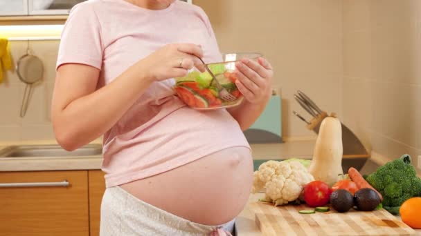 Primo piano della donna incinta con grande ventre contenente ciotola di vetro con insalata di verdure e mangiare in cucina — Video Stock
