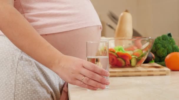 Primer plano de la mujer embarazada con el vientre grande de pie en la cocina y beber agua de vidrio. Concepto de estilo de vida saludable, nutrición e hidratación durante el embarazo — Vídeos de Stock