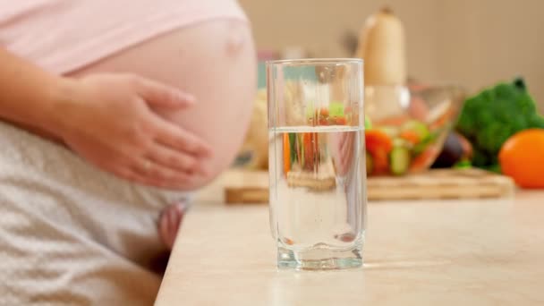 Hamile bir kadının mutfakta karnına dokunup okşamasına karşı dolu bir bardak su. Hamilelik boyunca sağlıklı yaşam biçimi, beslenme ve susuzluk kavramı — Stok video