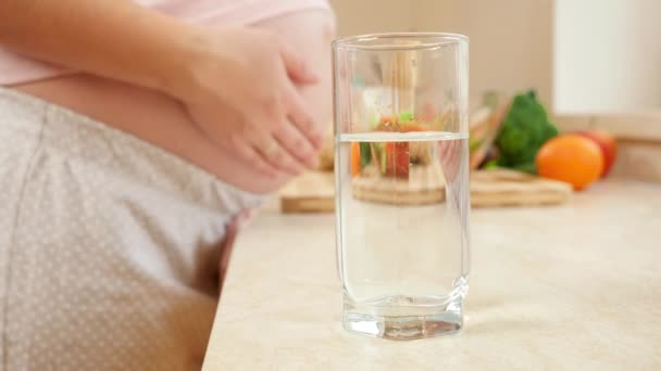 Primo piano della donna incinta che tocca la pancia grande e prende un bicchiere d'acqua dalla cima del tavolo della cucina. Concetto di stile di vita sano, nutrizione e idratazione durante la gravidanza — Video Stock