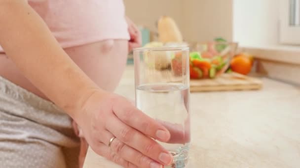 Крупним планом красива вагітна жінка бере склянку води і п'є її на кухні. Концепція здорового способу життя, харчування та гідратації під час вагітності — стокове відео