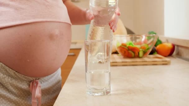 Primo piano di giovane donna incinta versando acqua in vetro e bevendola in cucina al mattino. Concetto di stile di vita sano, nutrizione e idratazione durante la gravidanza — Video Stock