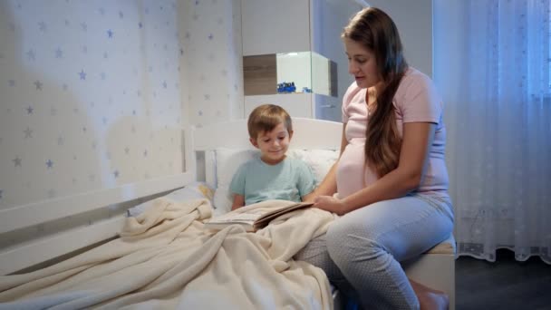 Mujer embarazada joven en pijama con su hijo leyendo un libro de cuentos de hadas por la noche antes de irse a dormir. Crianza y embarazo — Vídeos de Stock