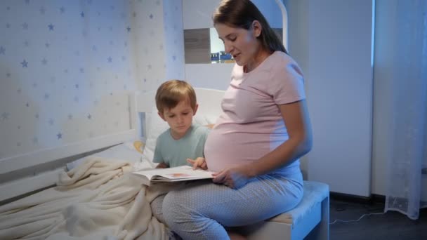 Улыбающийся мальчик с беременной матерью читает книжку в постели по ночам. Родители читают своим детям и проводят время вместе — стоковое видео