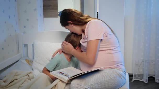 Молодая беременная мать обнимает и успокаивает своего плачущего маленького мальчика, лежащего ночью в постели. Заботливый и любящий родитель обнимает ребенка — стоковое видео