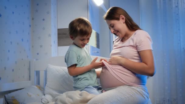 Ψαλίδι μικρό αγόρι ψάχνει και αγγίζει μεγάλη κοιλιά της εγκύου μητέρας του περιμένει το μωρό στο κρεβάτι το βράδυ — Αρχείο Βίντεο