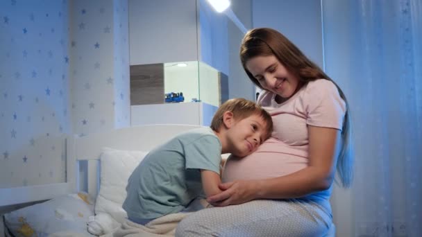 Sonriente niño pequeño abrazándose y hablando con el bebé nonato en el vientre grande si madre embarazada sentada al lado de su cama por la noche — Vídeos de Stock