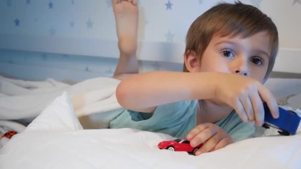 Lächelnder kleiner Junge, der nachts im Bett mit zwei Spielzeugautos spielt. CHild spielt vor dem Schlafengehen. — Stockvideo