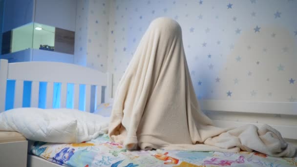 Happy child jumping out and uncovering big blanket at bedroom at night. Funny active toddler boy palying and scaring at night — Stock Video