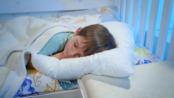 Panning video malého chlapce v pyžamu spánek na posteli pod bílou přikrývkou v noci. Dítě odpočívá a má v noci dobré sny — Stock video