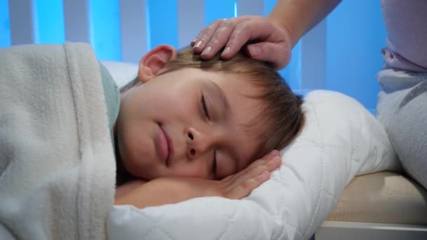 Närbild av omtänksam mor sitter på sängen bredvid sovande liten pojke på natten och smeker honom på huvudet med handen — Stockvideo
