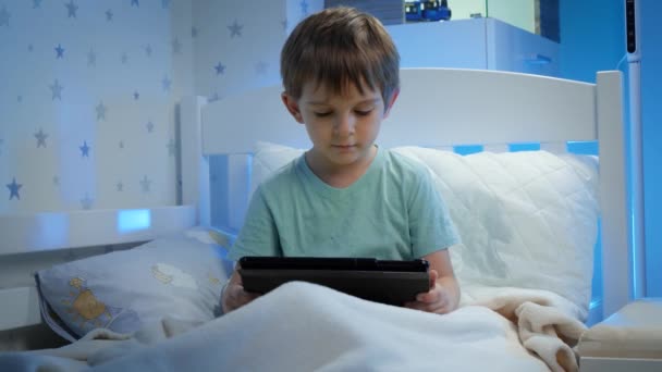 Retrato de menino pequeno sentado na cama à noite e assistindo vídeo no computador tablet. Menino com gadget à noite — Vídeo de Stock