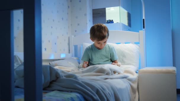 Mały 5-letni chłopiec korzystający i bawiący się na tablecie przed pójściem spać w nocy. Dziecko z gadżetem wieczorem — Wideo stockowe