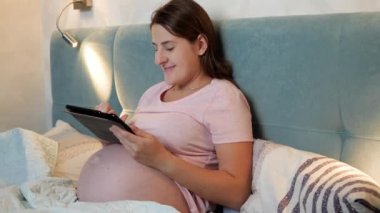 Koca göbekli, gülümseyen hamile bir kadının portresi. Yatağa yatmadan önce tablet bilgisayar kullanıyor..