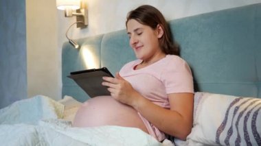 Pijama giyen, tabletle çalışan ve geceleri yatmadan önce internette gezinen gülümseyen hamile bir kadın.